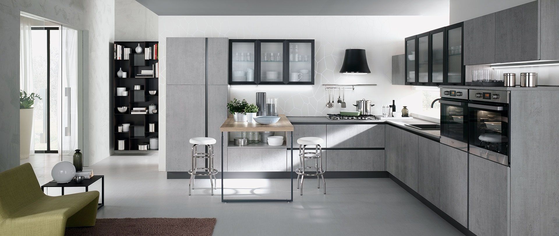 Moderna cucina grigia con isola, forno e mobili neri. È visibile una zona soggiorno con una sedia verde.