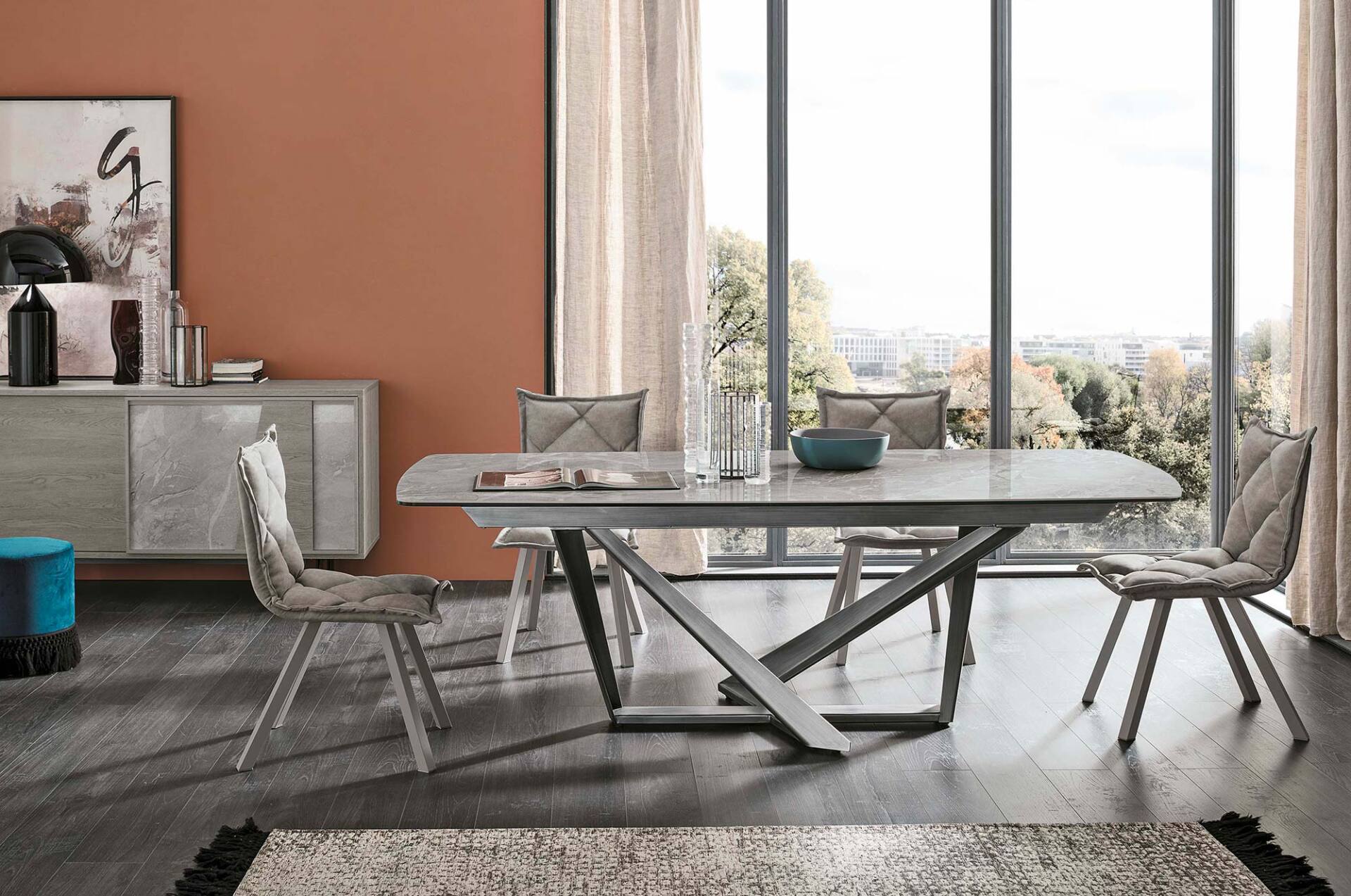 Sala da pranzo con tavolo, sedie, credenza, tappeto e ampie finestre con vista. Arredamento nei toni della terra.