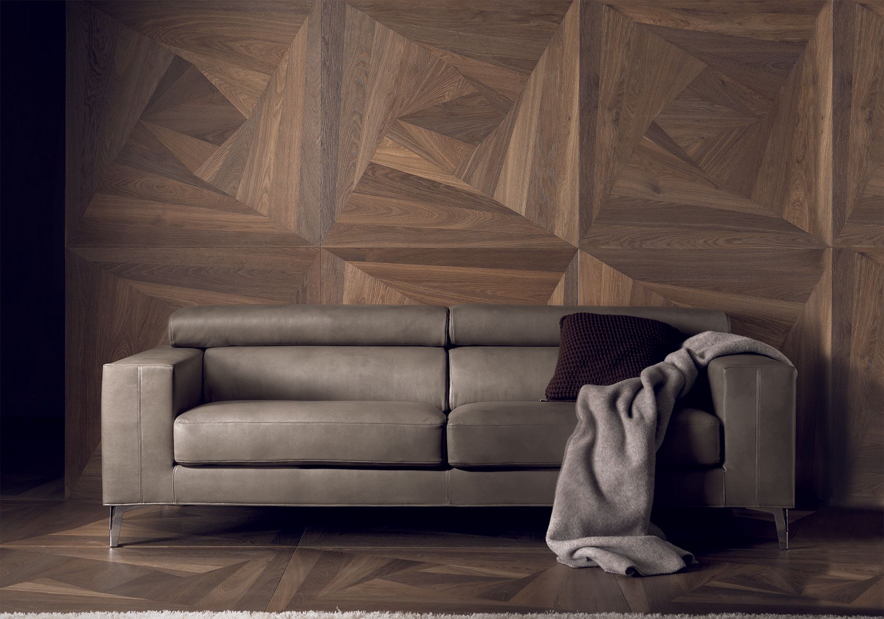 Divano in pelle grigia contro una parete geometrica in legno, con coperta e cuscino.