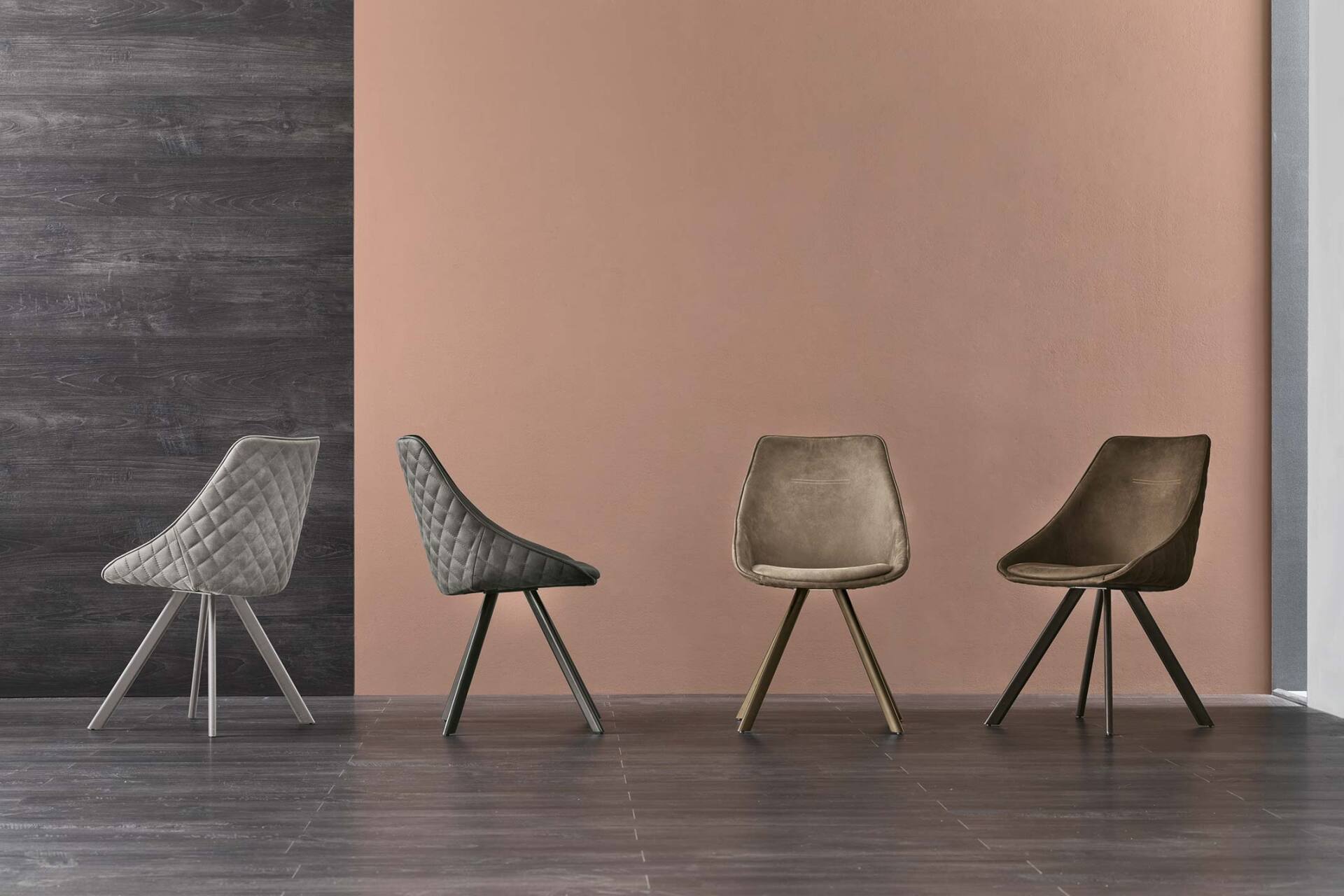Quattro sedie moderne con gambe triangolari, di colori e texture diversi, contro pareti strutturate.