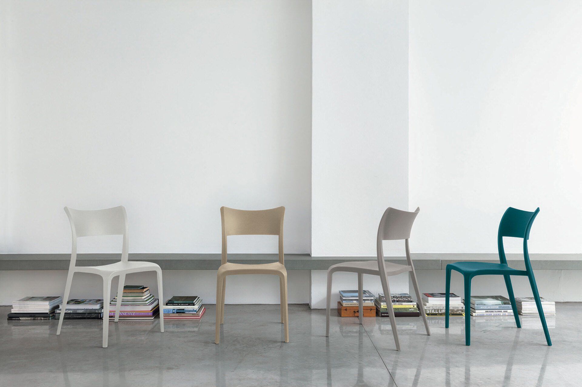 Quattro sedie di plastica color bianco sporco, beige, grigio e verde acqua contro una parete bianca; sotto di esse ci sono dei libri.