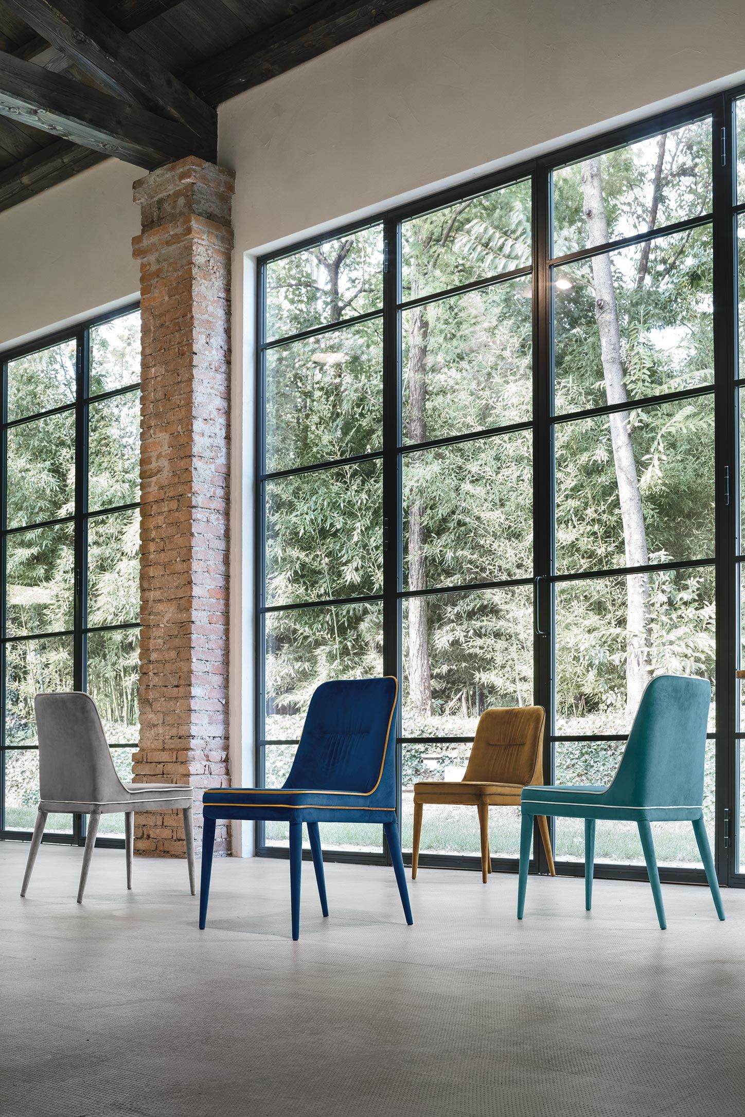 Quattro sedie moderne in una stanza con grandi finestre, colori vari: grigio, blu, senape, verde acqua.