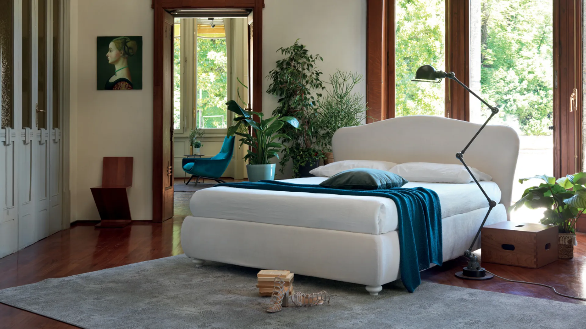 Camera da letto con letto bianco, tappeto grigio e lampada alta. Piante verdi e ampie finestre con luce naturale.