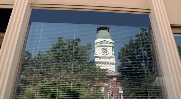 A clock tower is reflected in a window with blinds