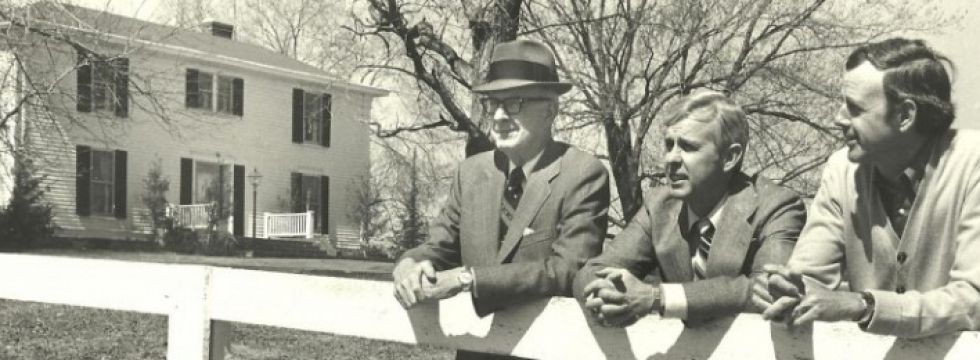 Three men are sitting on a fence in front of a house.