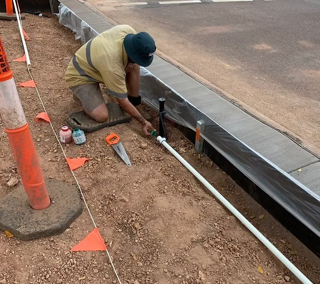 A construction worker kneels, cutting a white pipe next to a curb. Orange cones and flags mark the work area. — iWater NT Pty Ltd in Yarrawonga, NT