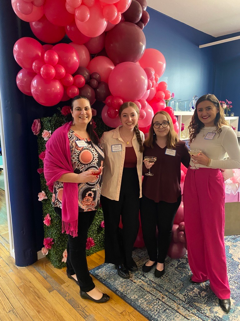 A group of women are posing for a picture in front of a wall of pink balloons.