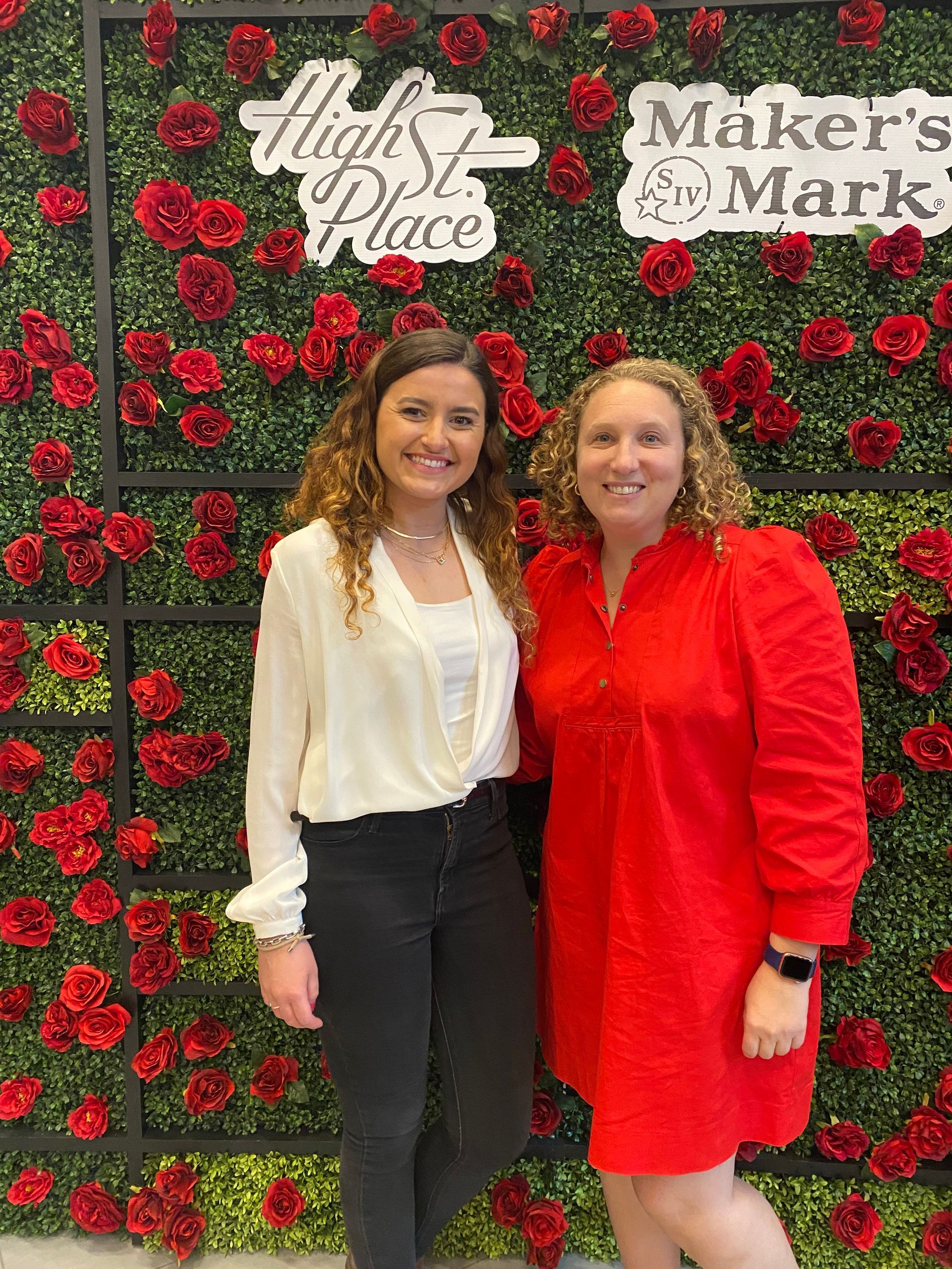 Two women are posing for a picture in front of a wall of red roses.