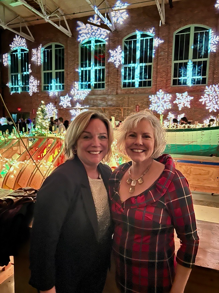 Two women are posing for a picture in front of a building with christmas lights on the windows.
