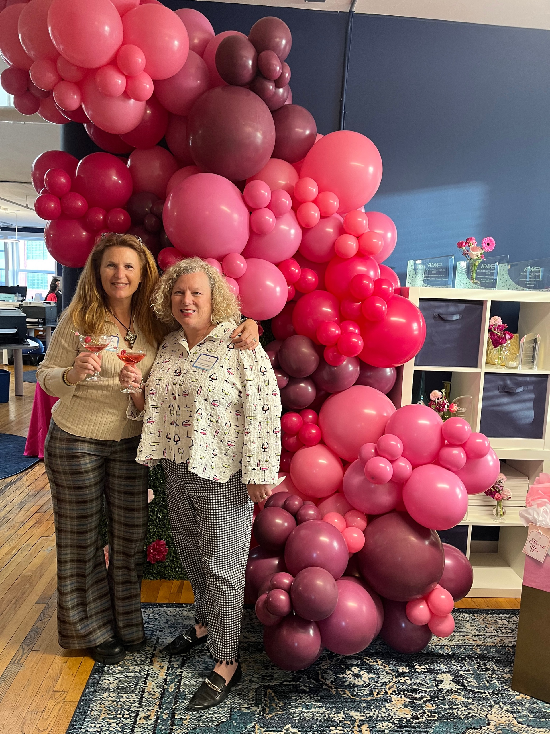 Two women are standing in front of a wall of pink balloons.