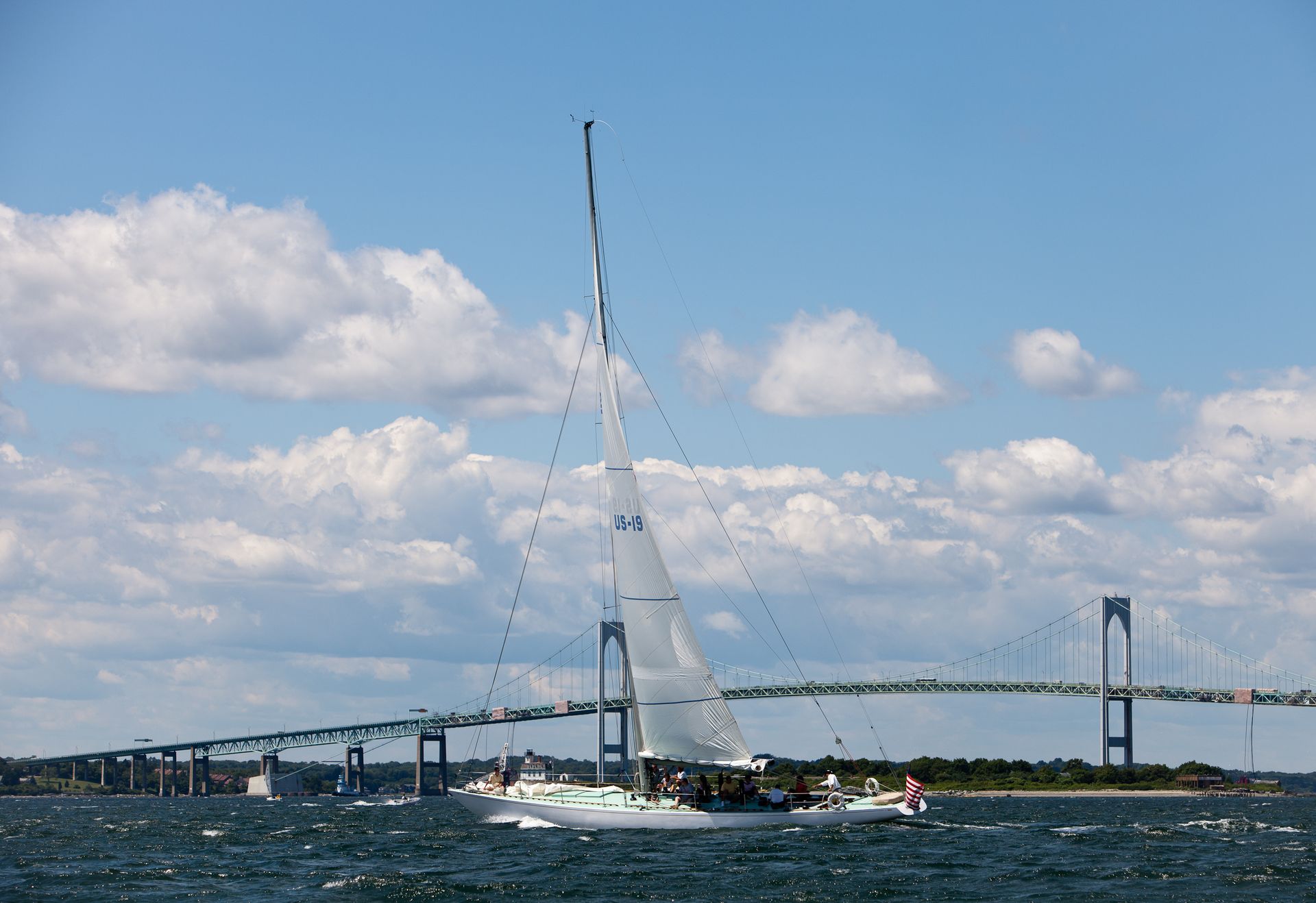 Sailboat sailing on water with a bridge in the background under a blue sky with fluffy clouds.