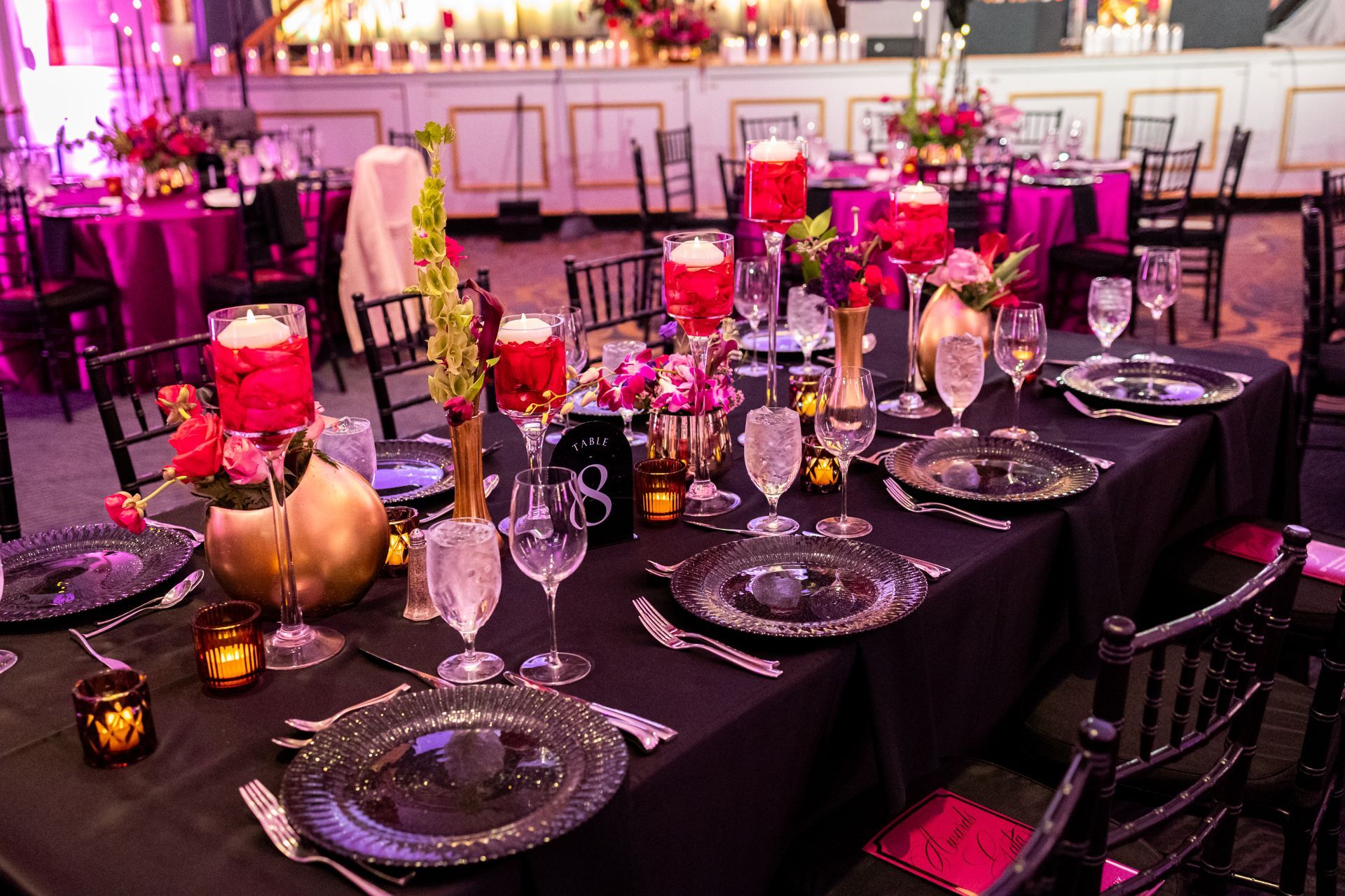Elegant banquet table with black linens, pink floral centerpieces, candlelight, and glassware in a decorated event hall