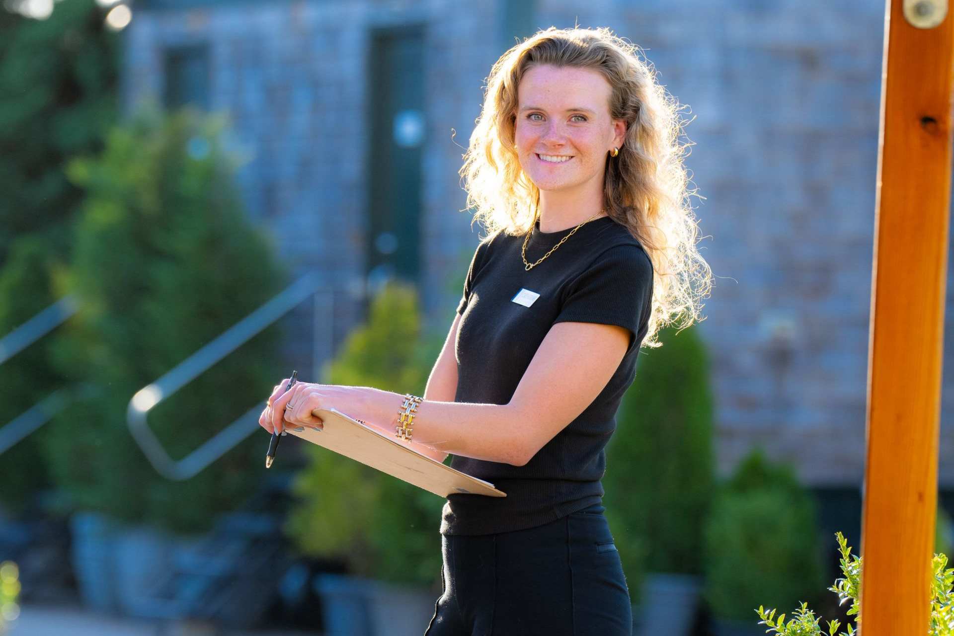 Woman in black shirt holding a clipboard outdoors, smiling in front of shrubbery and a wooden structure.