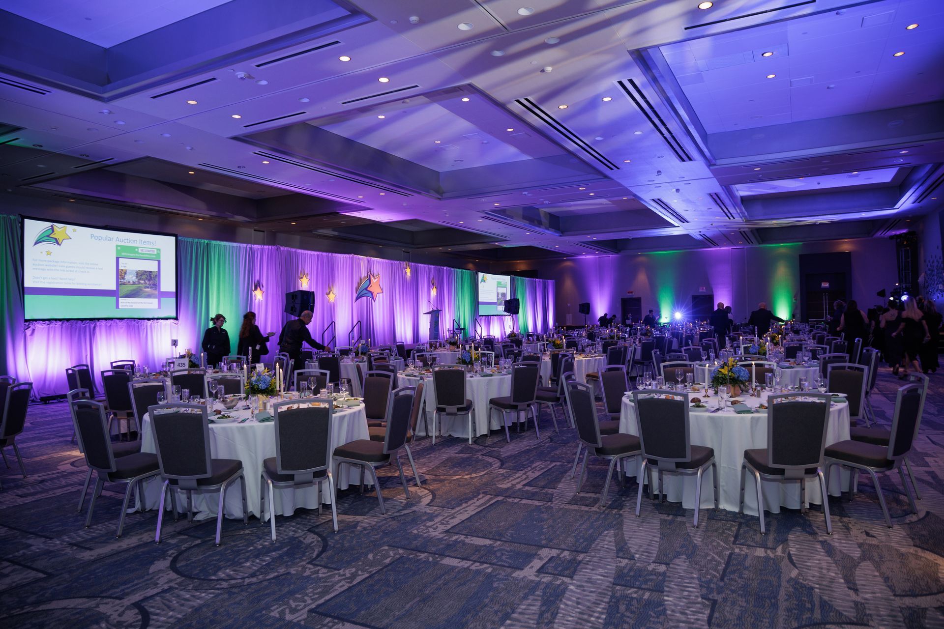 Elegant banquet hall with round tables, chairs, and screens, illuminated by purple and green lighting.