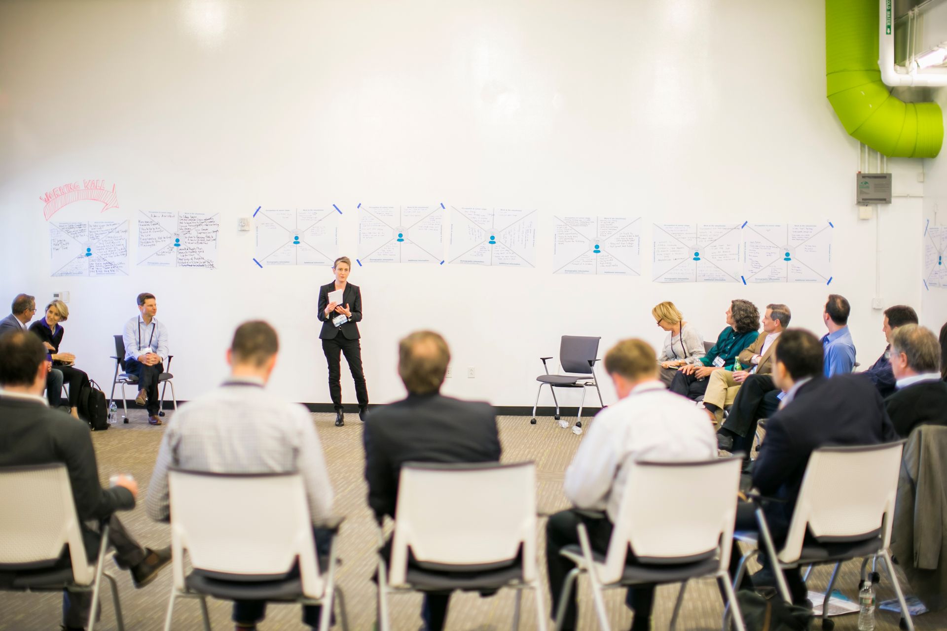 Woman presenting in front of a group of people sitting in a circle; white walls with drawings.