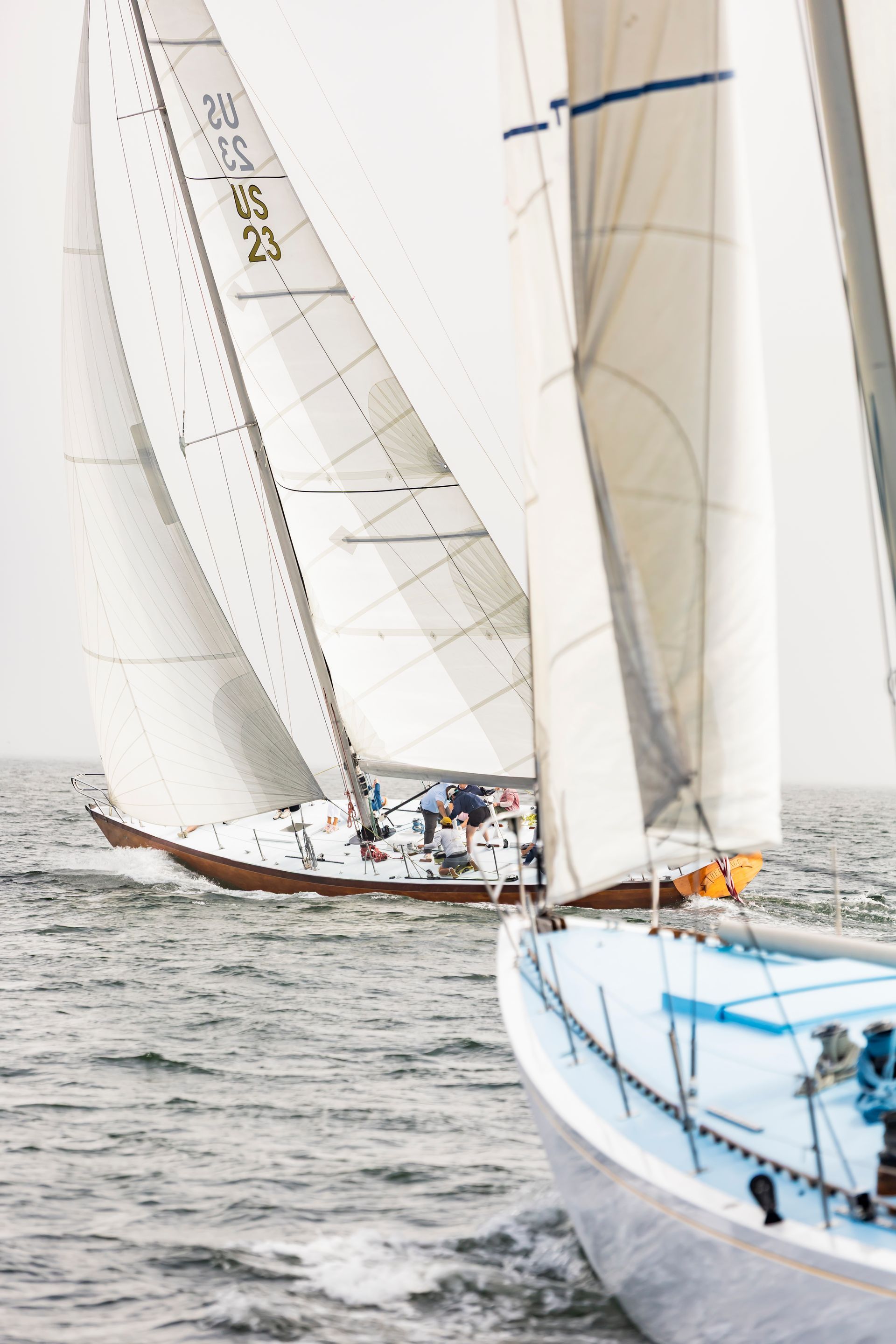 Two sailboats racing on a gray, choppy sea. The lead boat is white and blue; the other is wood-toned.