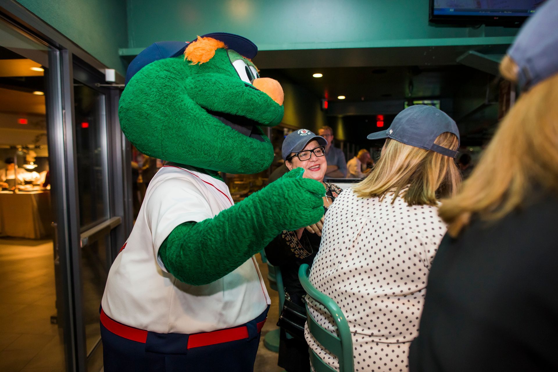 Green baseball mascot giving a thumbs-up to a woman wearing a blue hat, in a restaurant.