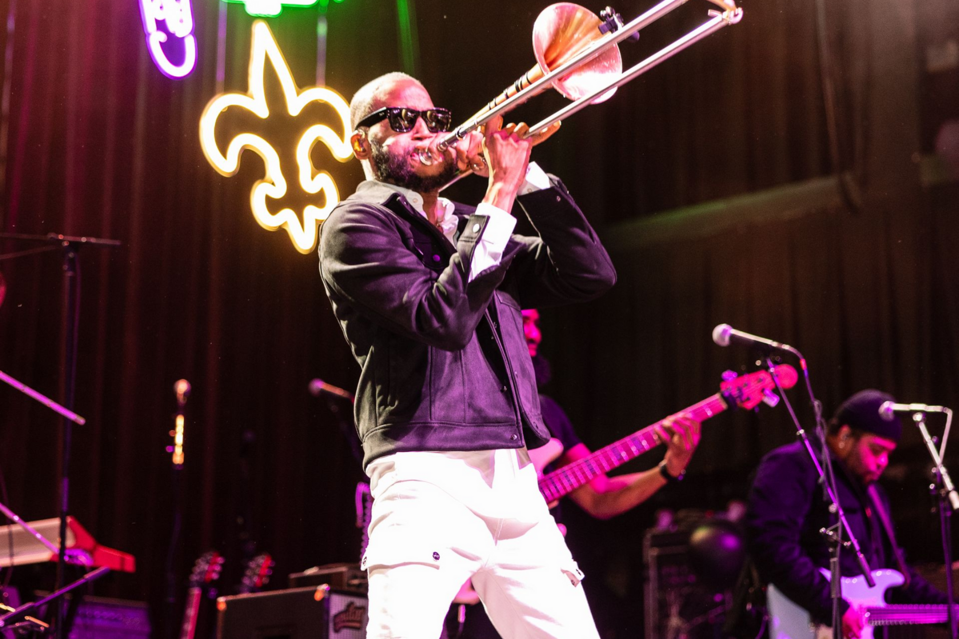 Man playing trombone onstage with a band, lit by stage lights and a neon sign.
