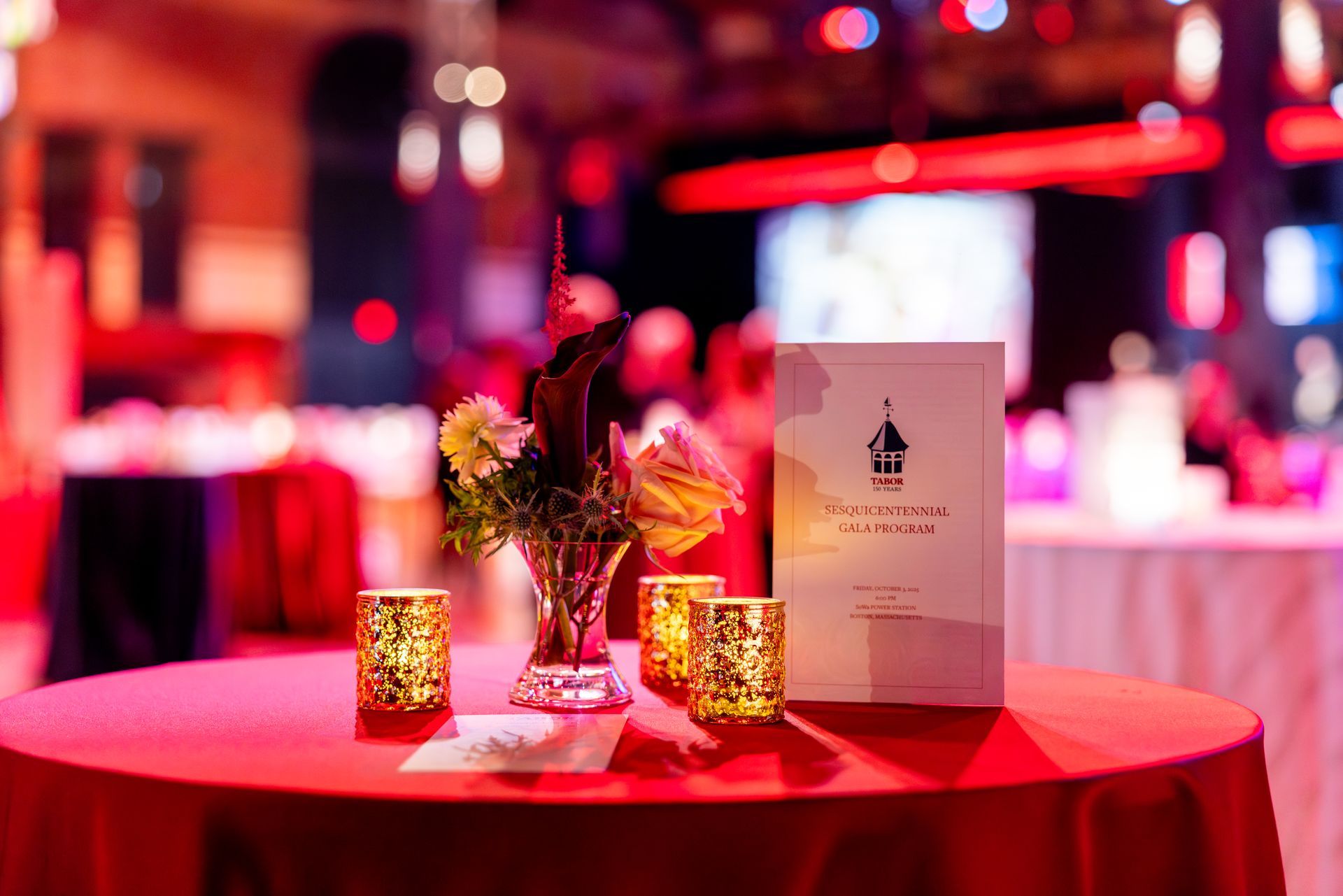 Red tablecloth with flowers, candles, and event sign in a festive, blurred setting.
