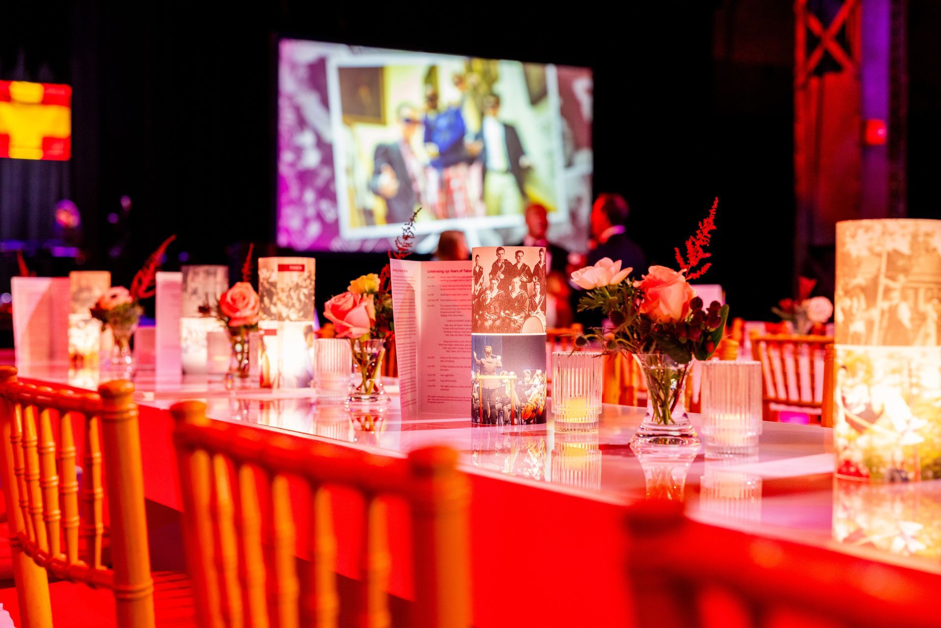 Long table set for an event with floral centerpieces, candles, and a large screen in the background.