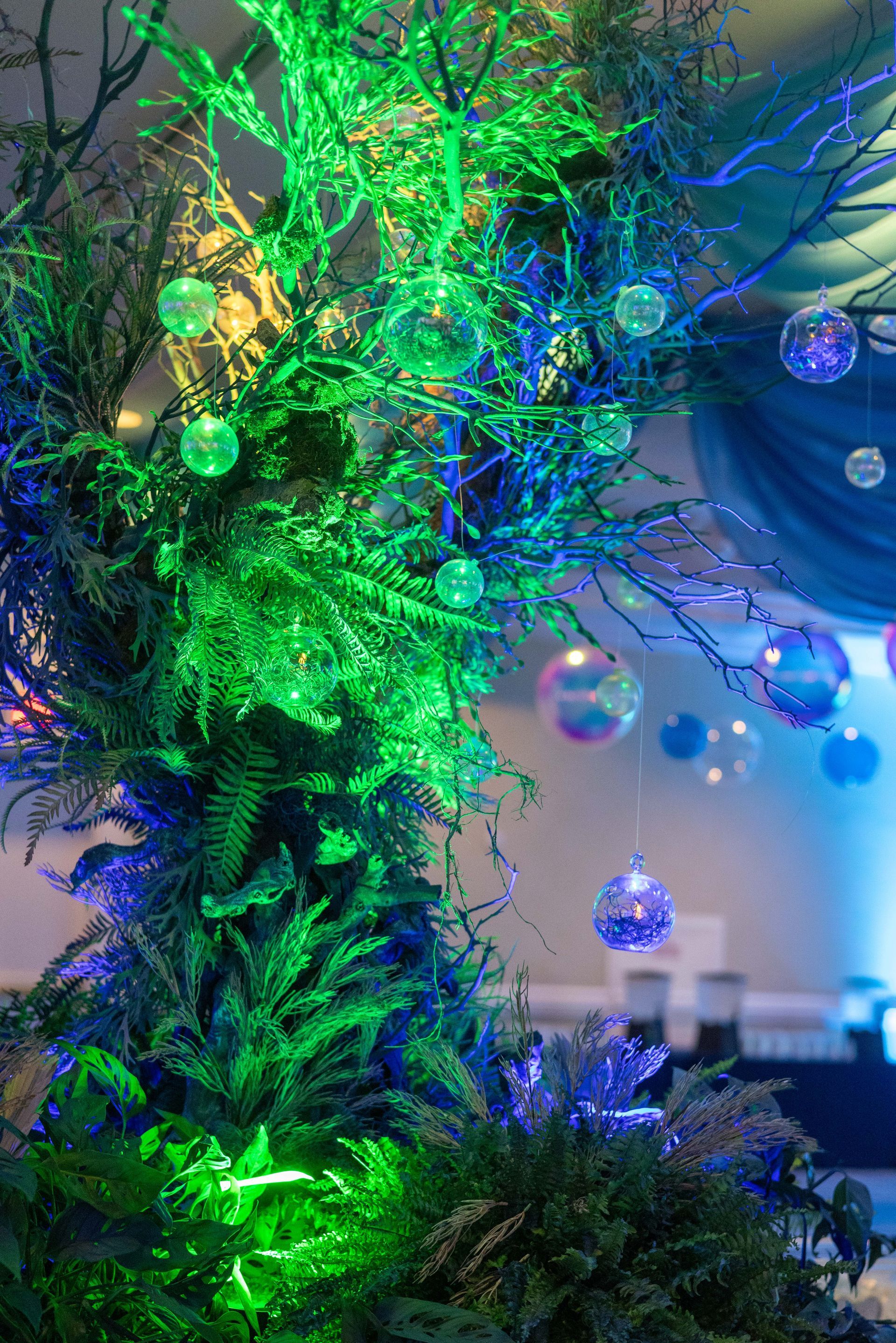Green and blue illuminated tree decorated with hanging iridescent orbs, indoors.