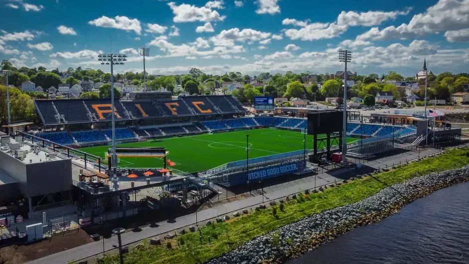Aerial view of a soccer stadium: Rhode Island FC. Blue seats, green field, and black scoreboard. Near a body of water.