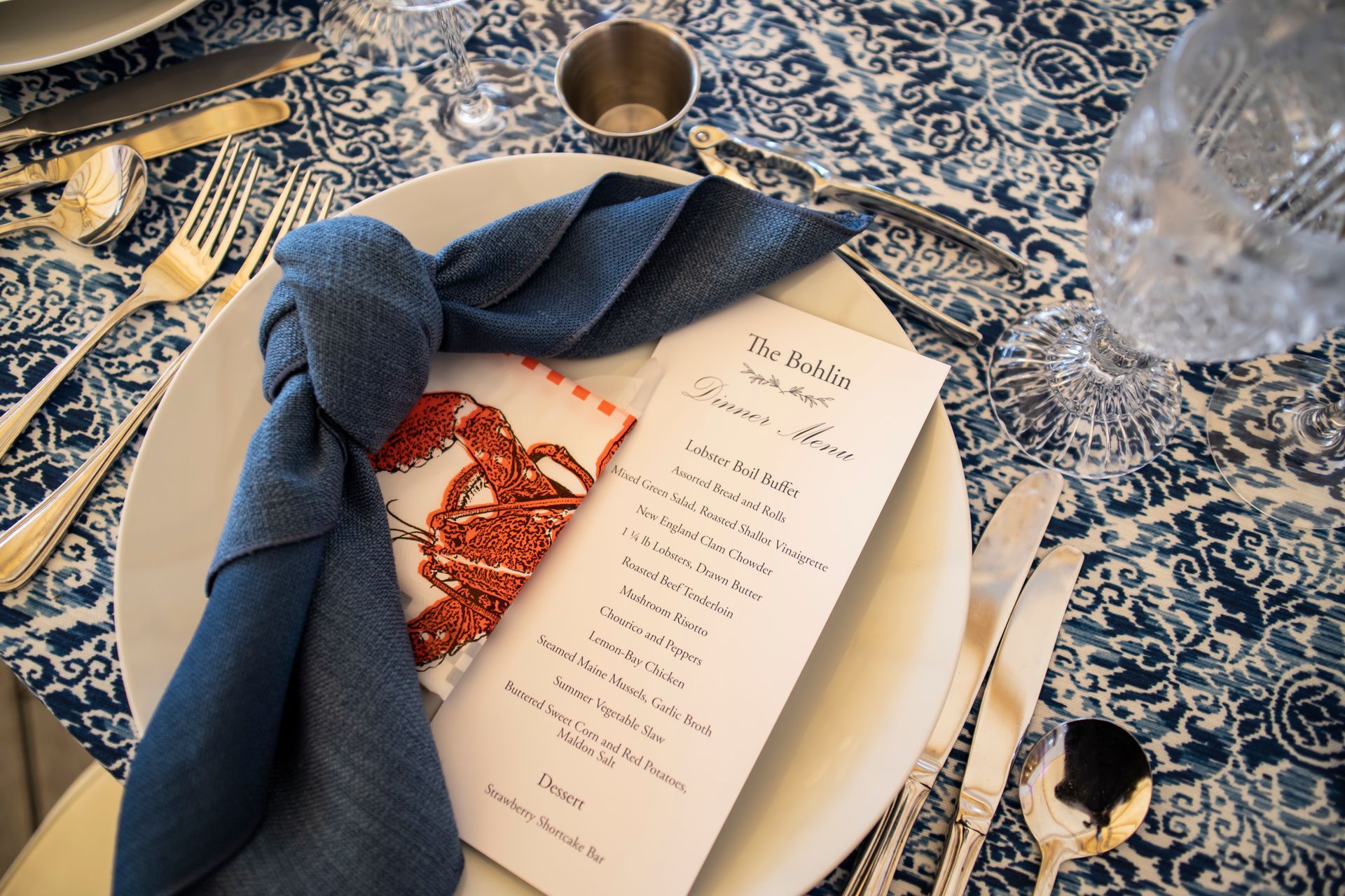 Place setting with blue-knotted napkin, menu on plate with silver cutlery, and blue patterned tablecloth.