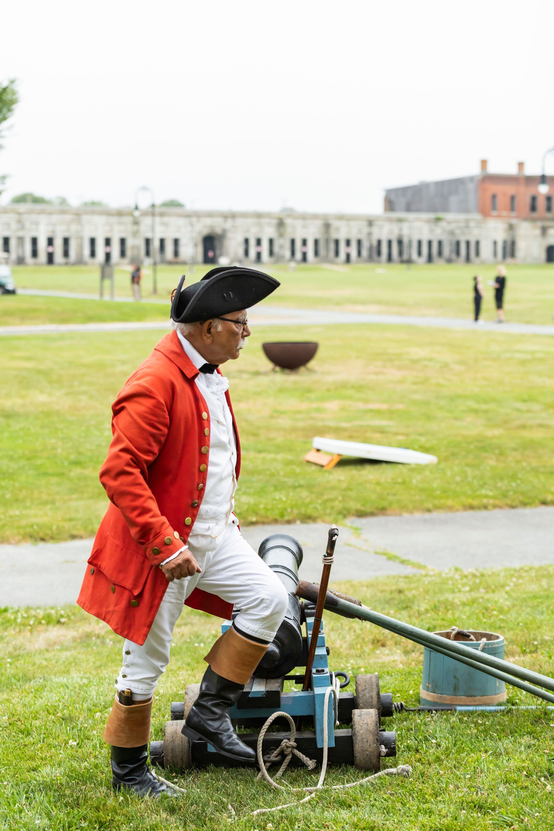 Man in red coat and tricorn hat, standing by a cannon on a green field, brick building in background.