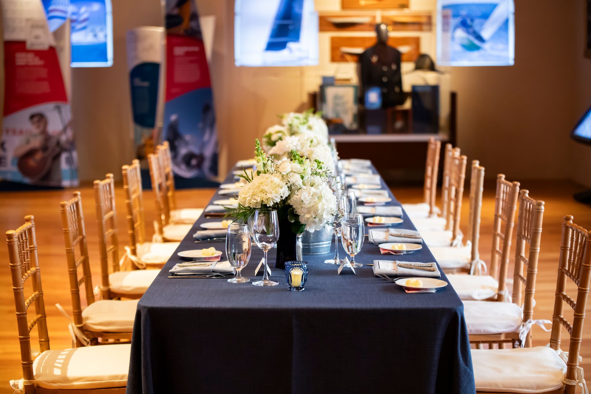 Long, formal banquet table with a dark blue tablecloth, white floral centerpieces, and gold chairs in a gallery setting.