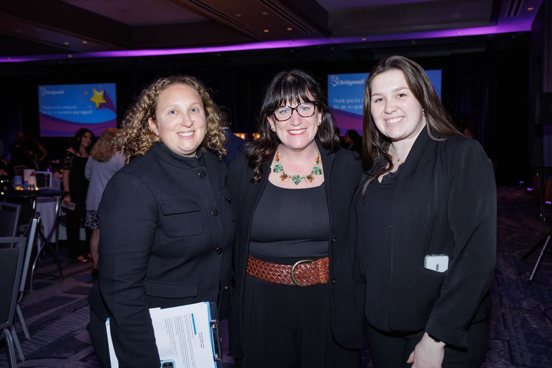 Three women smiling, posing at an event. One in center with glasses, brown belt, others in black jackets. Purple lighting.