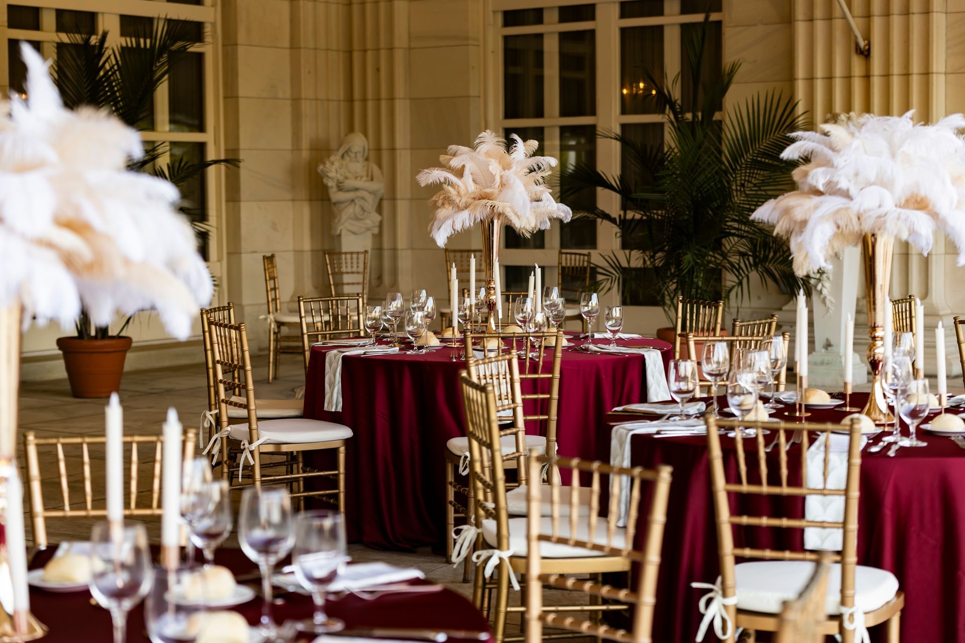 Elegant outdoor dining tables set for a formal event, burgundy tablecloths, gold chairs, feathered centerpieces.