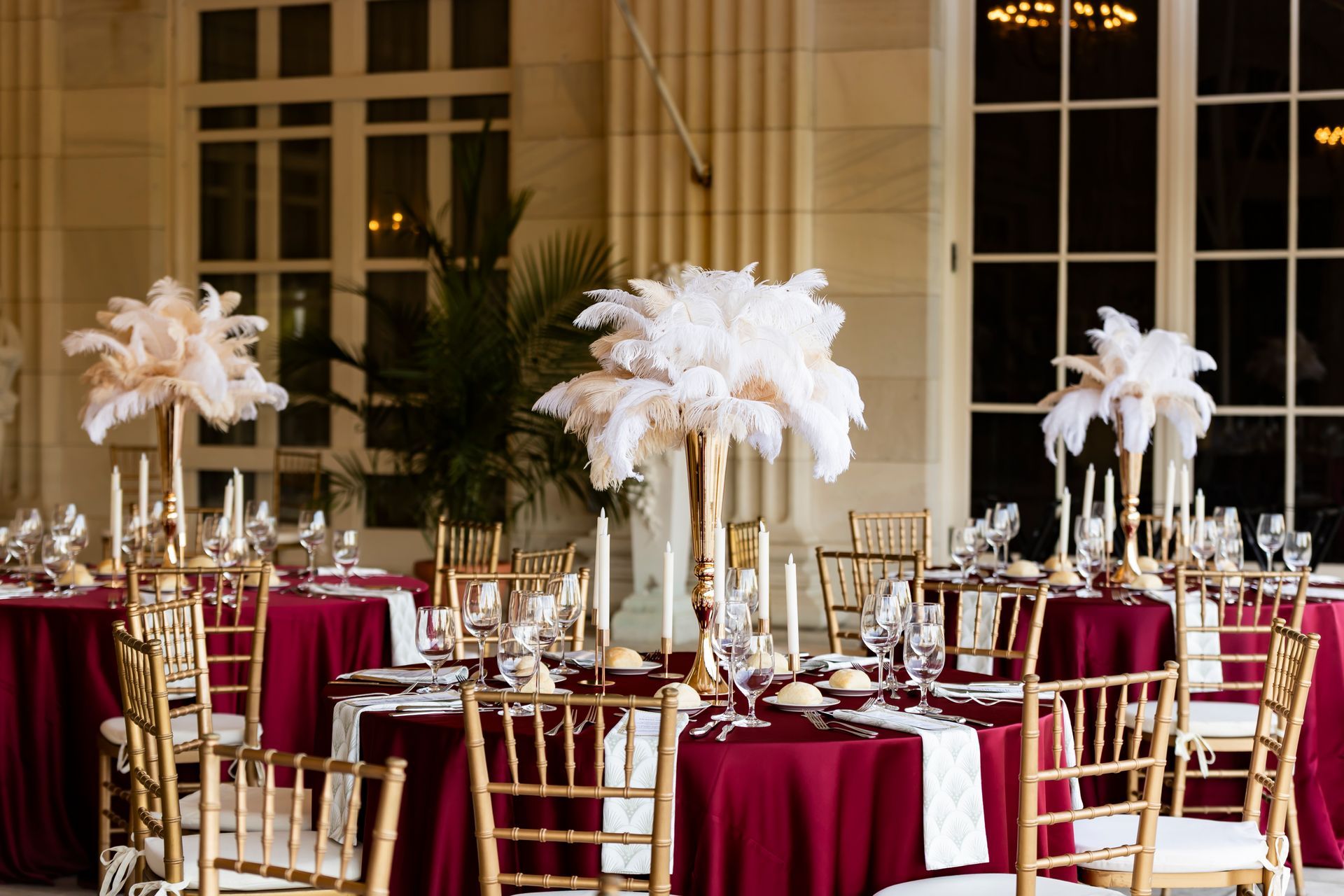 Round tables with burgundy tablecloths, gold chairs, and tall ostrich feather centerpieces in a bright event space.