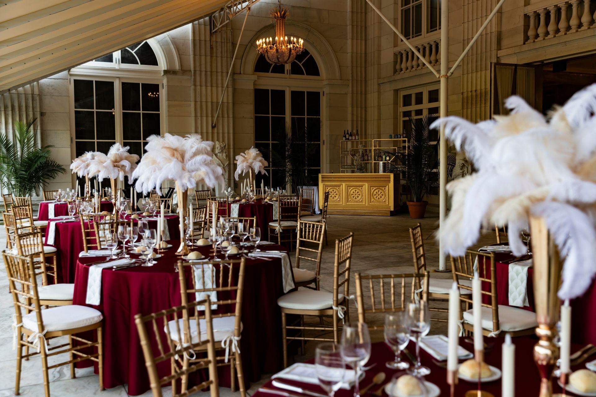 Formal banquet setup with burgundy tablecloths, gold chairs, and feather centerpieces.