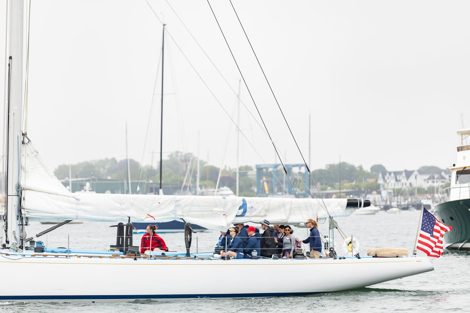 A white sailboat cruises on a harbor with a crew on deck and an American flag flying at the stern.