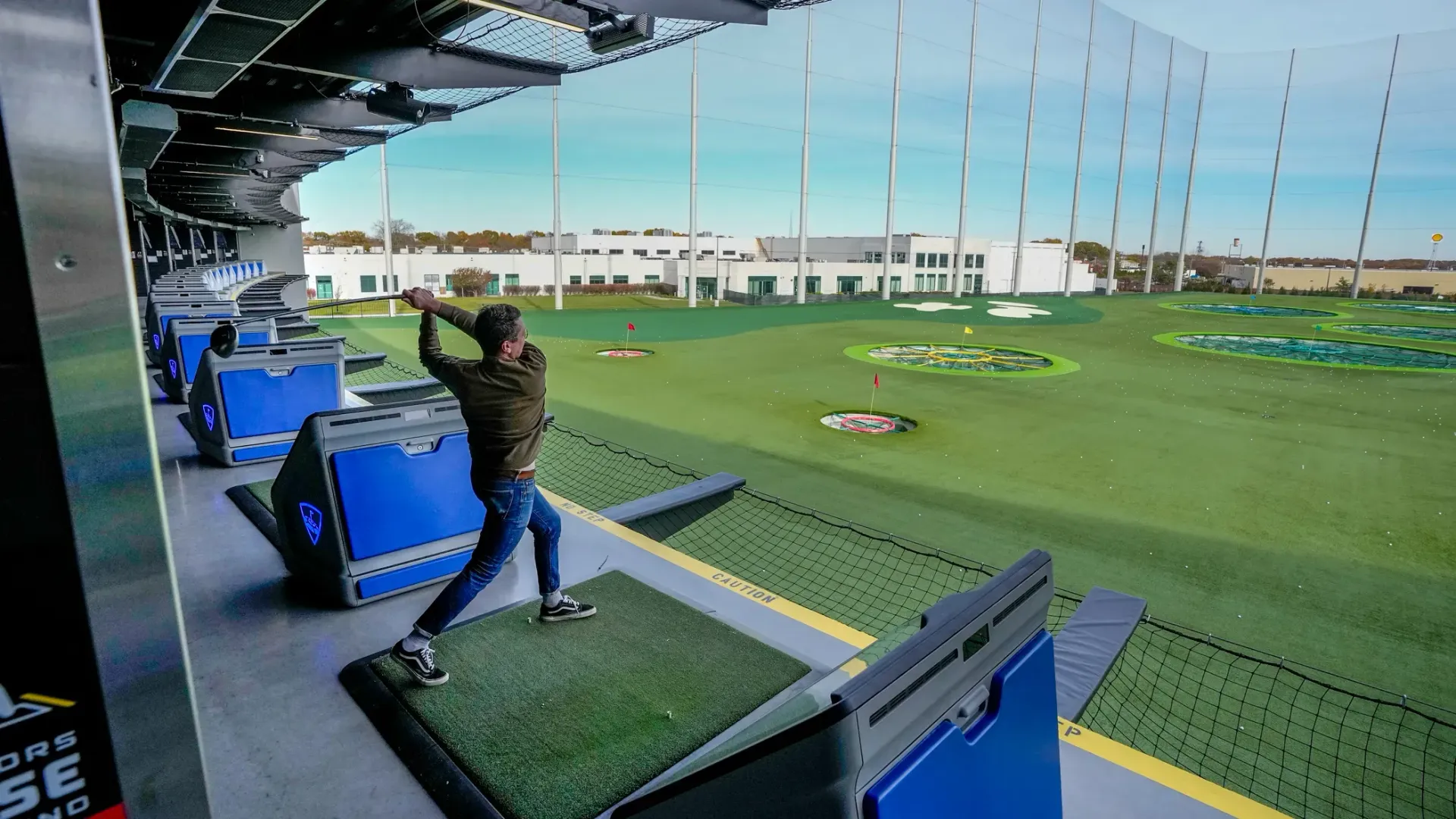 Person swinging a golf club at a Topgolf, wide shot. Blue bays, green grass, targets, and netting visible.