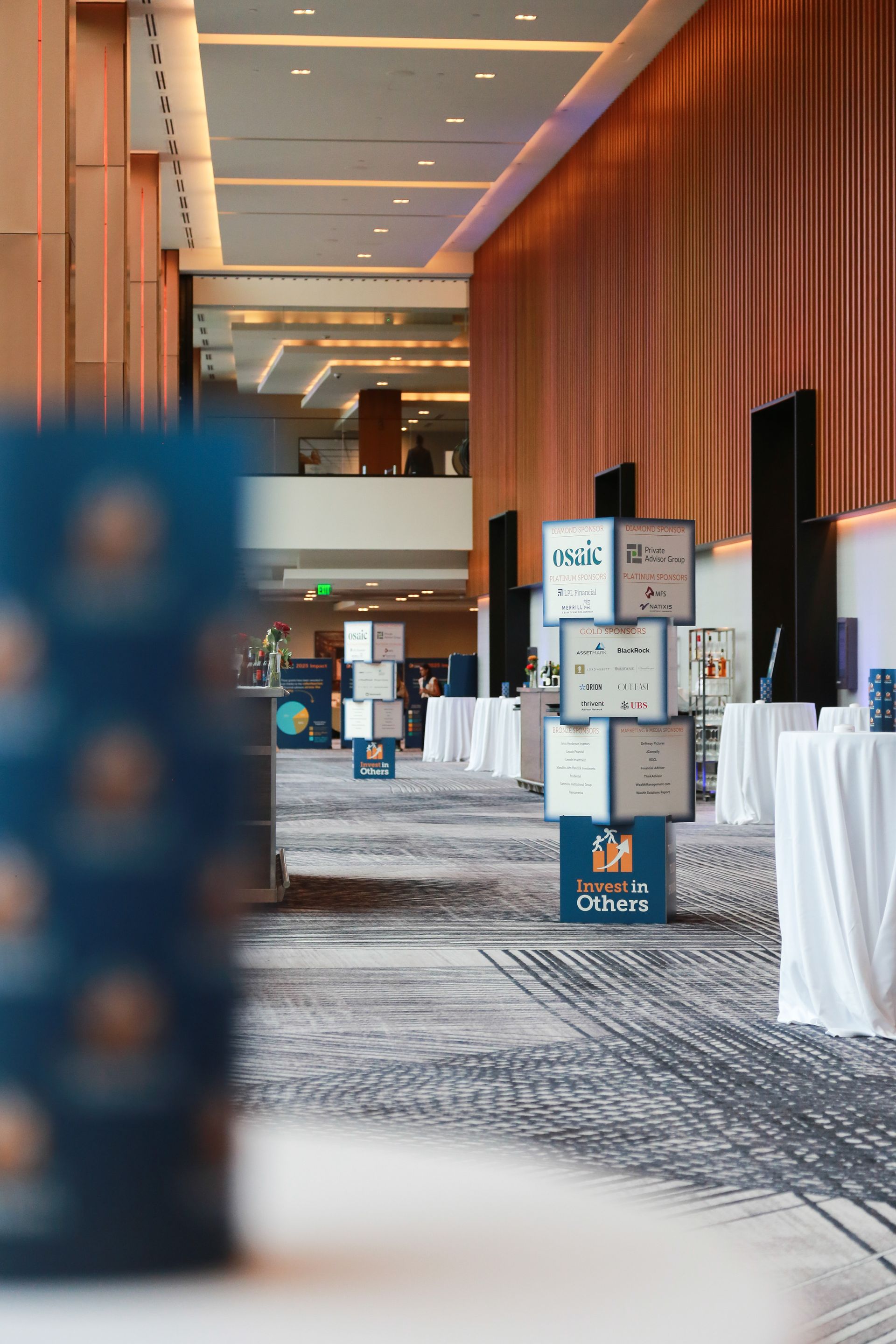 Hallway with event signage and white tablecloth-covered tables. Modern decor with light wood paneling, carpet.