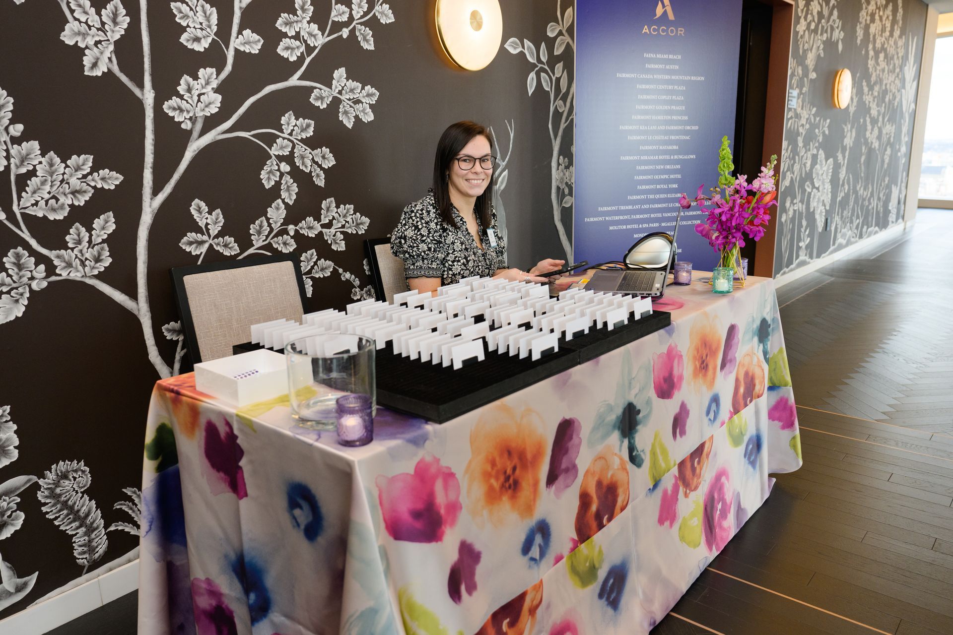 Woman behind a table with jewelry samples, floral tablecloth, and patterned wall.