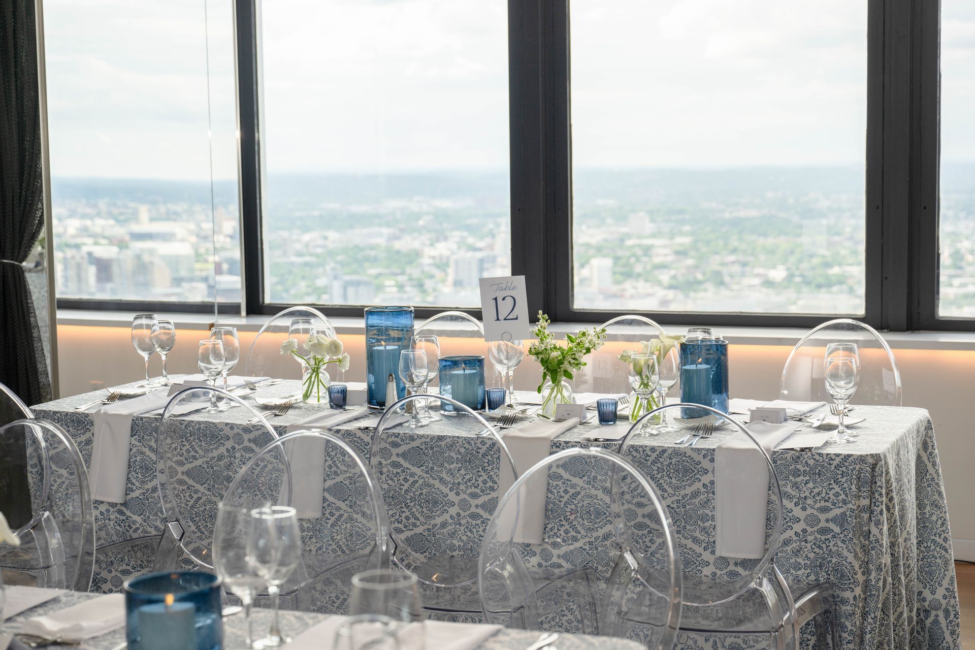 Table setting with city view; blue and white linens, clear chairs, and centerpieces with flowers and candles.