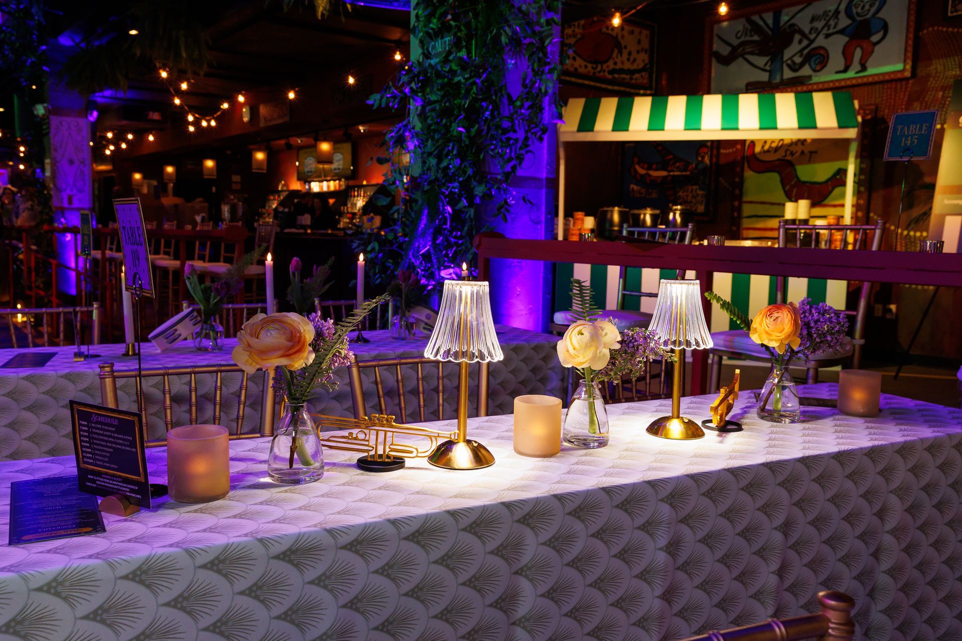 Long table set for a gathering, featuring lamps, candles, flowers, and a patterned tablecloth. Dimly lit, festive setting.