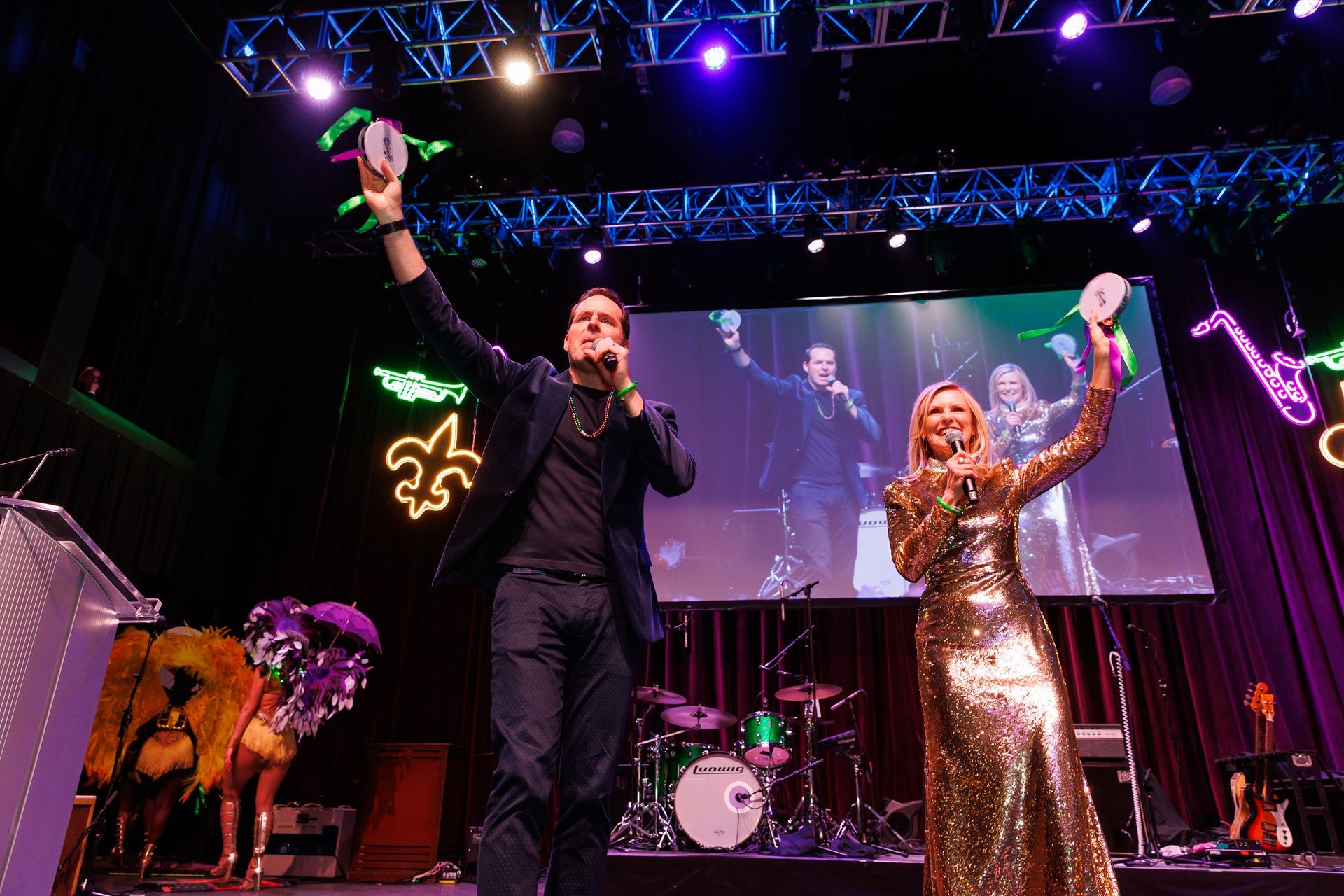 Man and woman on stage holding masks, smiling, with stage decorations and a screen in the background.