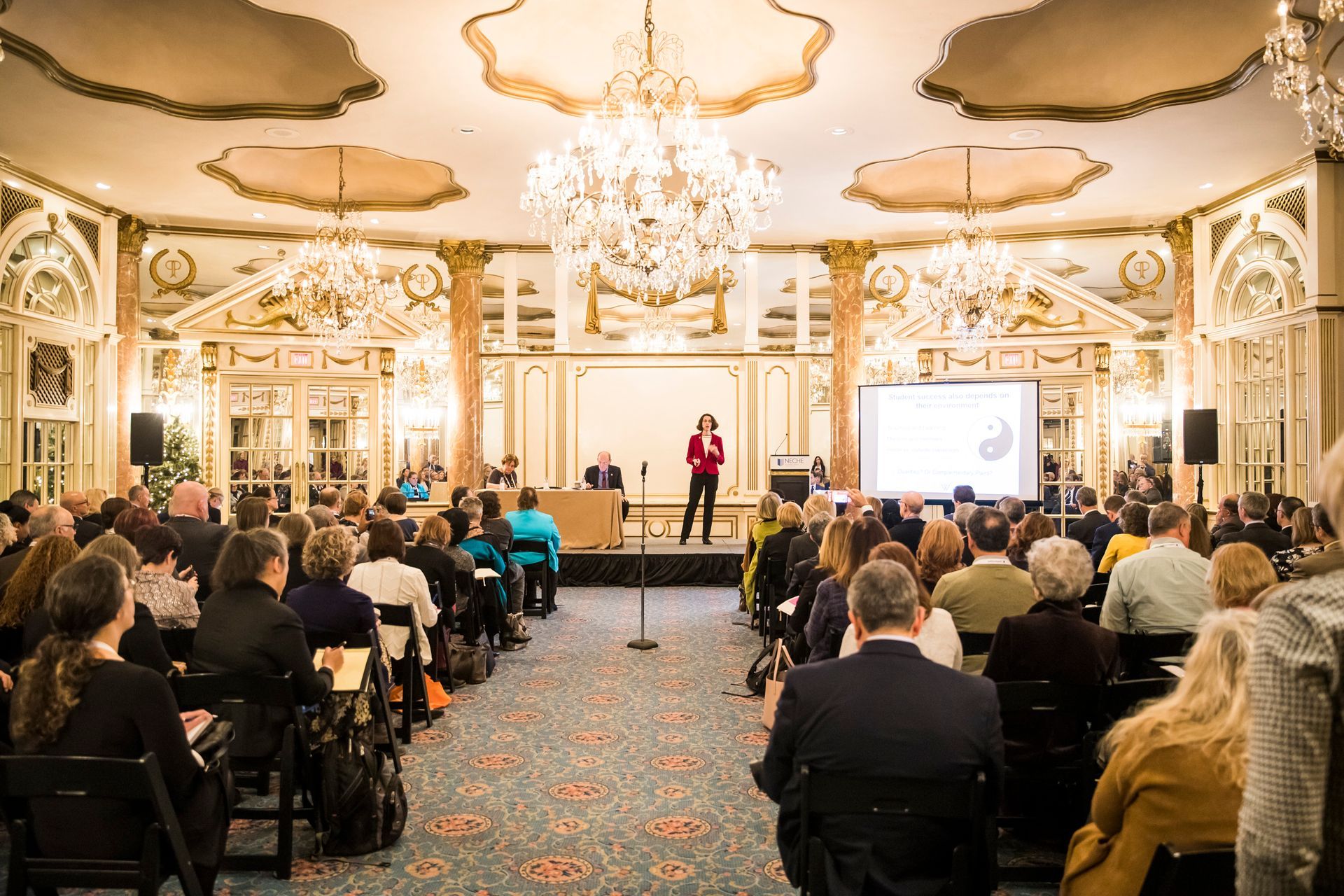 A woman presents at a conference in a grand ballroom with an audience, lit by a chandelier.