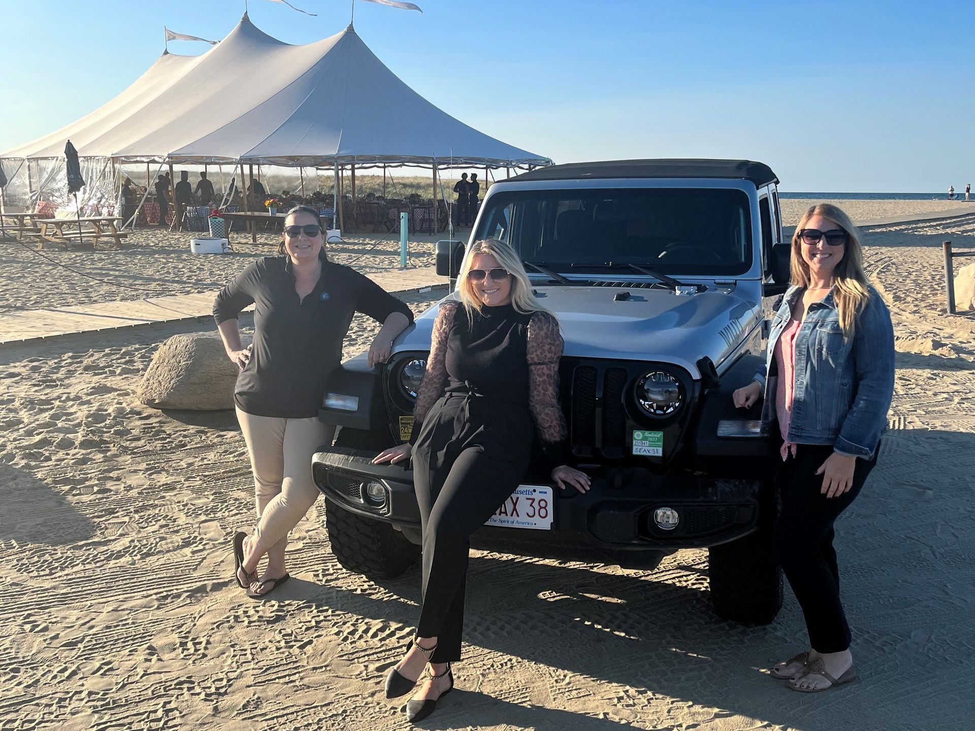 Three women pose with a black Jeep on a beach, a white tent in the background.