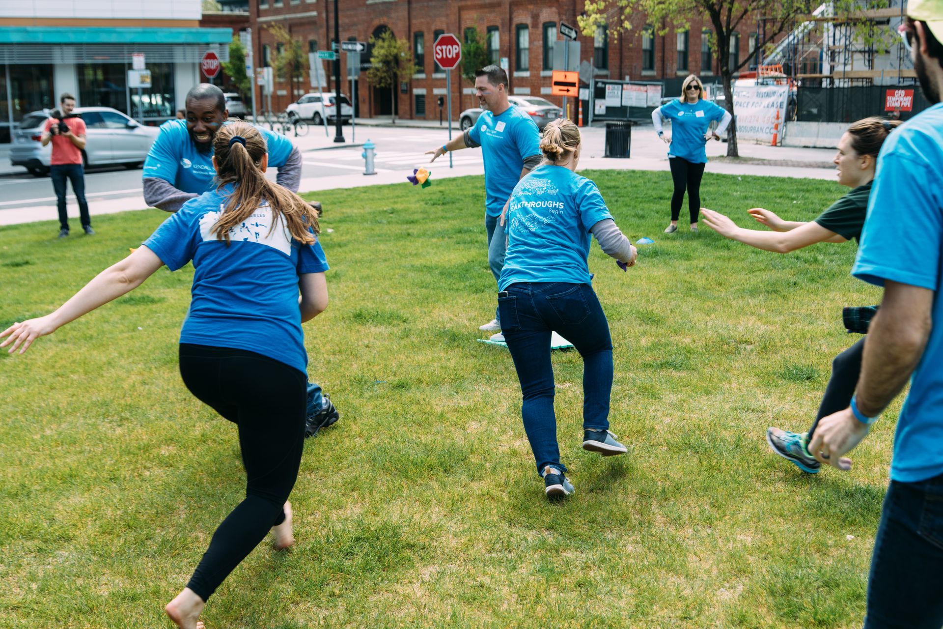 People playing a game on a grassy area in an urban setting. Most are wearing blue shirts and are running or reaching.