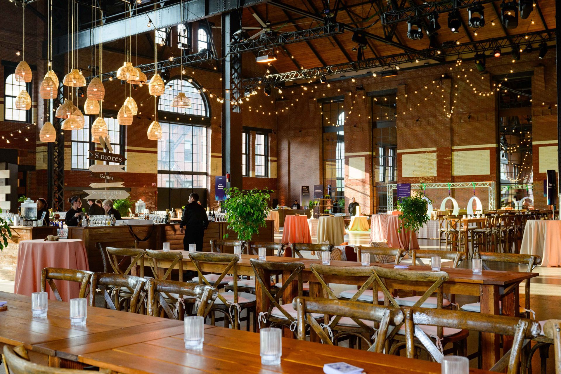 Event space with long wooden tables, decorated with lights and plants, brick walls.