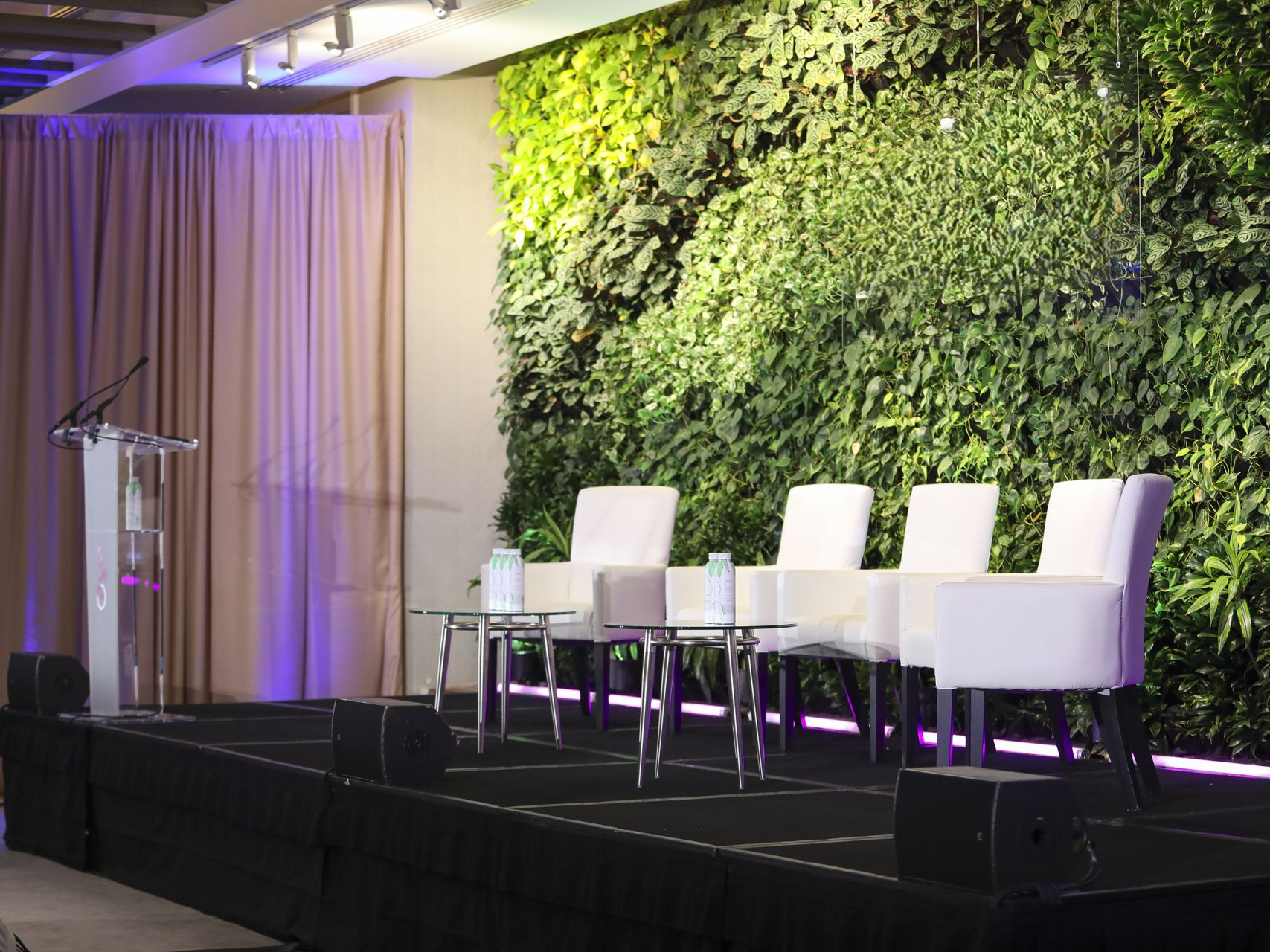 A stage with white chairs, glass tables, and a podium. A green living wall provides the backdrop.