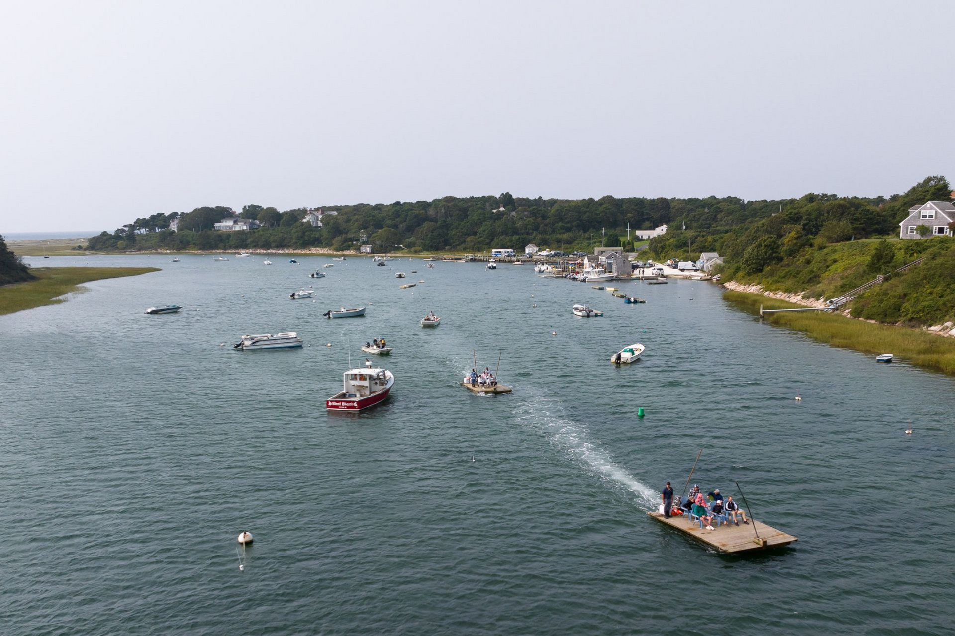 Boats on a calm, blue waterway; a large dock with people; green trees and buildings in the background.