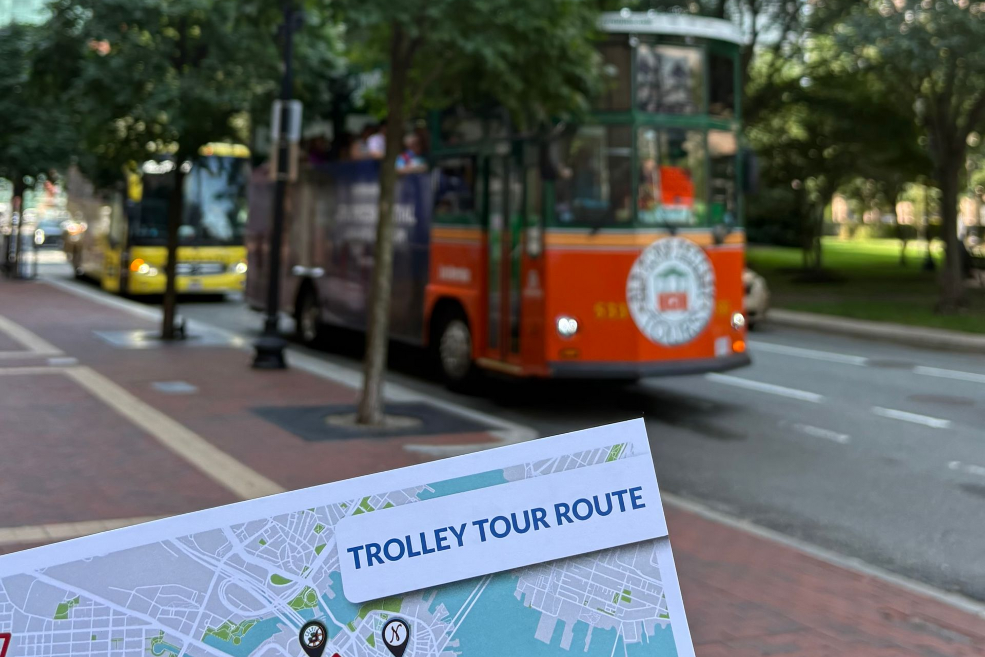 Trolley tour route map in focus, with an orange trolley and yellow bus in the background on a city street.