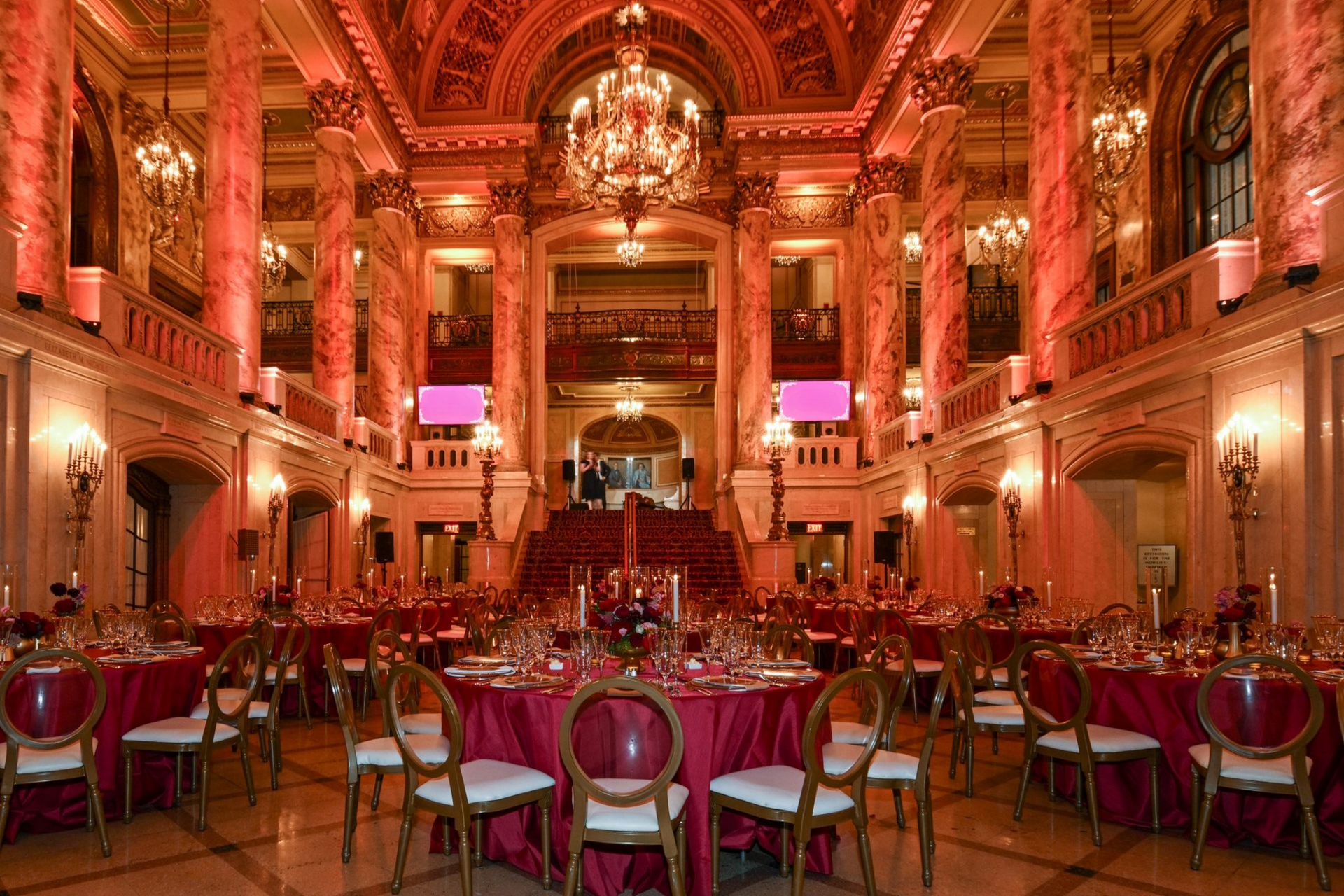 Formal dining hall with round tables, burgundy tablecloths, and ornate architecture. Crimson lighting illuminates the space.