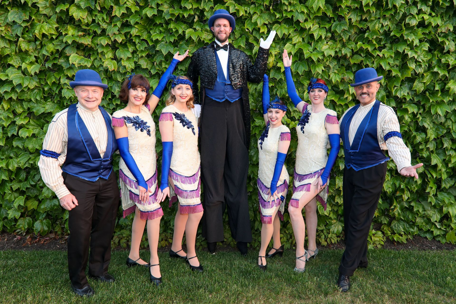 Group of seven people in vintage-style costumes pose in front of a green wall; the tallest man in the center.