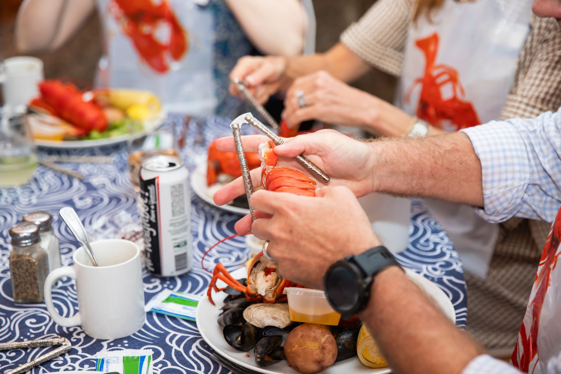 People cracking lobster claws at a table set for a seafood meal, with bibs, food, and drinks visible.